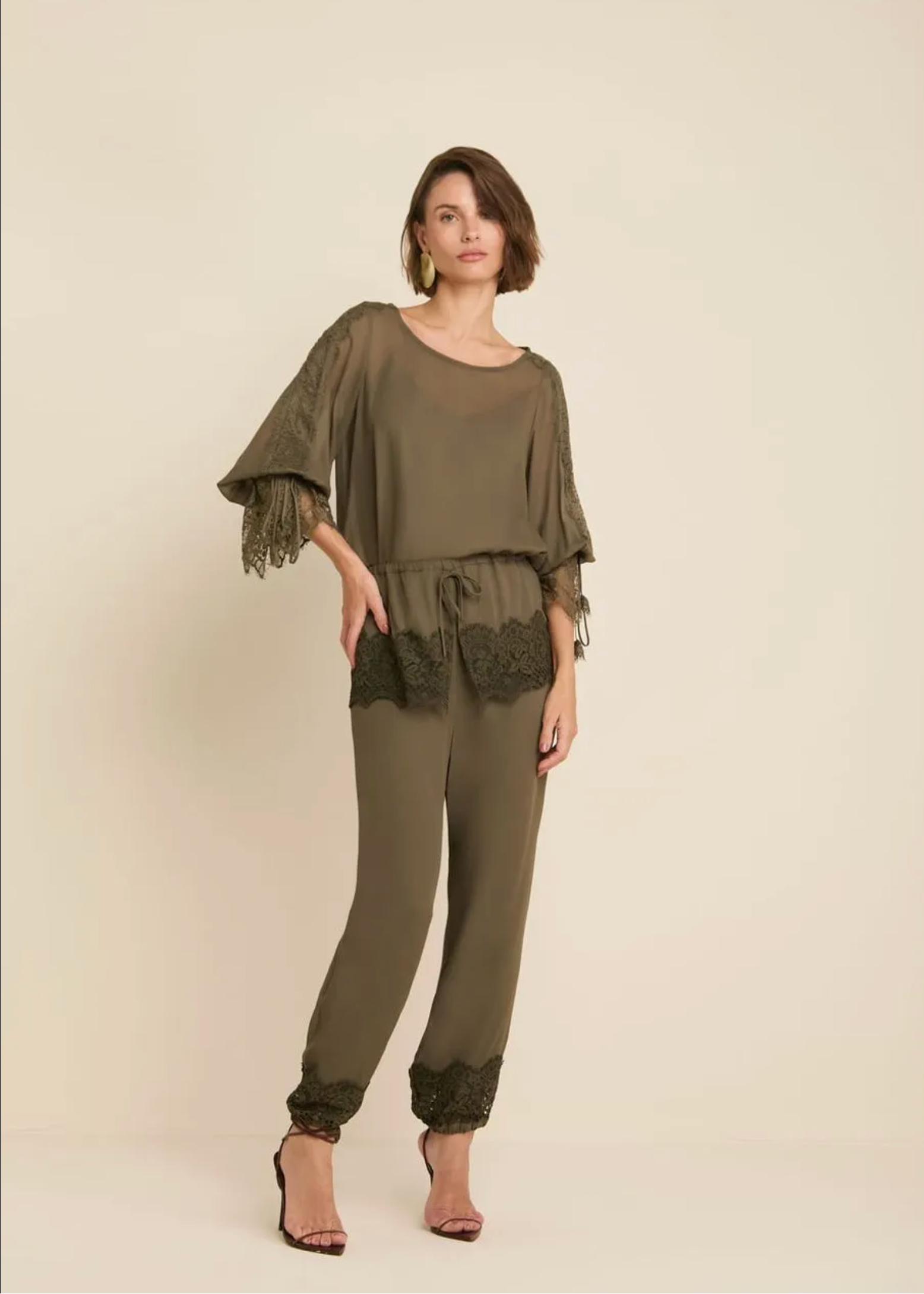 A woman modeling an olive green two-piece jumpsuit with lace trim details, consisting of a loose short-sleeve top with lace flutter sleeves and matching lace-trimmed pants, set against a plain beige studio background.