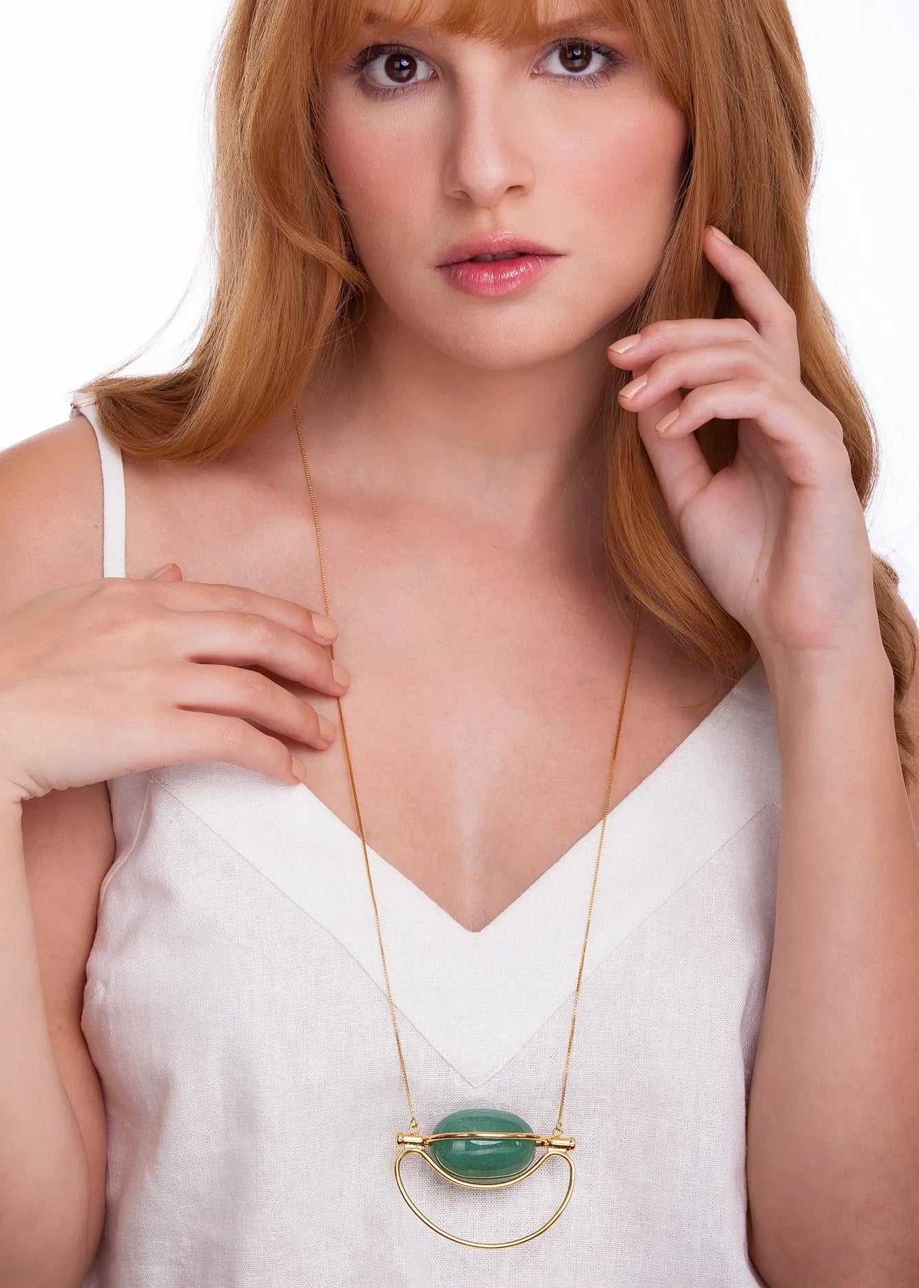 A woman with red hair wearing a white camisole top and a gold pendant necklace with a green oval stone, photographed against a plain white background.