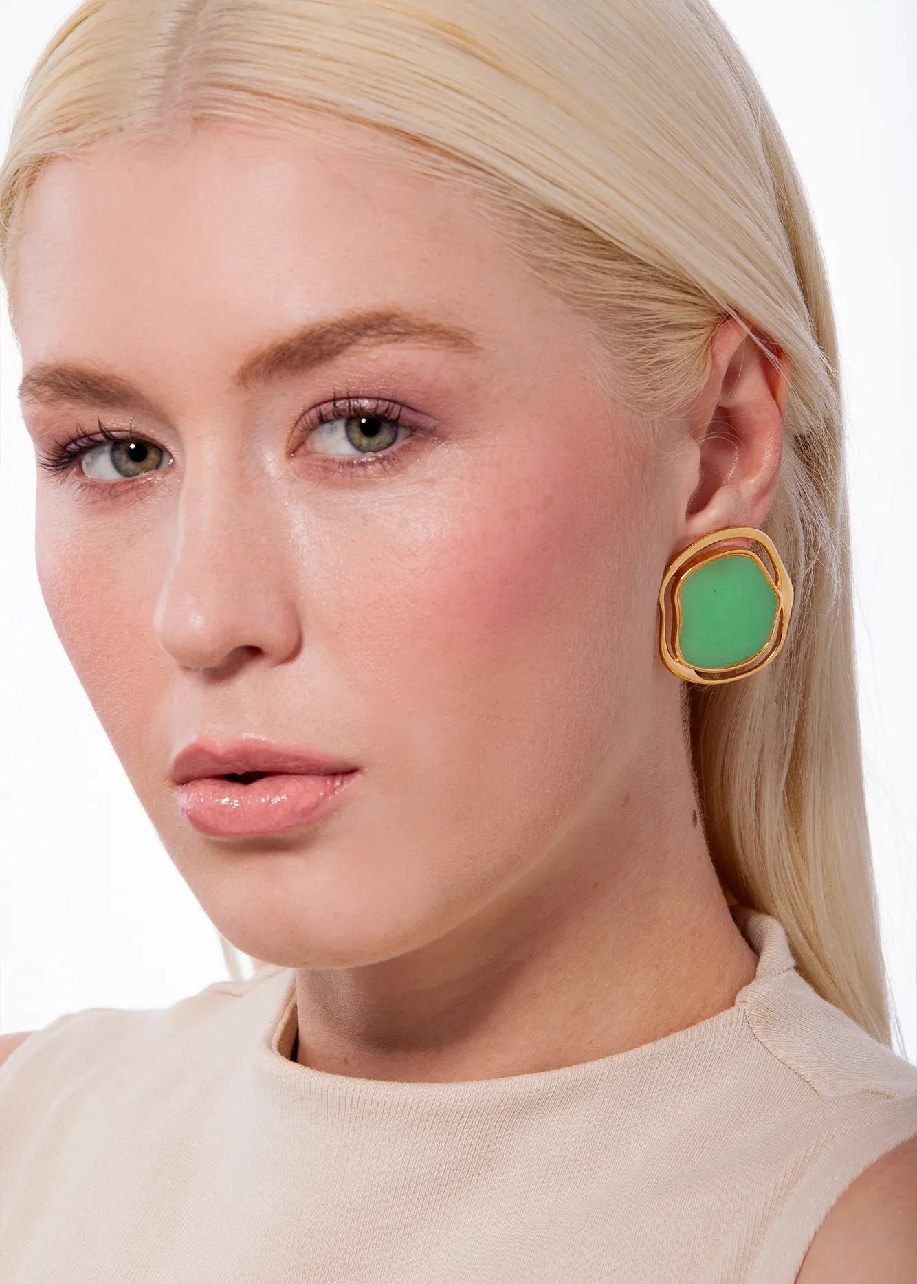 A close-up of a blonde woman wearing a chunky gold earring with a green stone, against a clean white background.