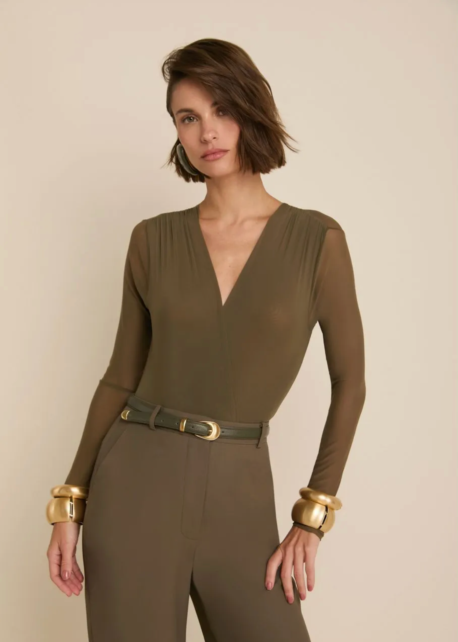 A woman modeling a solid olive green long-sleeve jumpsuit with a matching belt and gold-tone cuffs, set against a plain beige studio background.