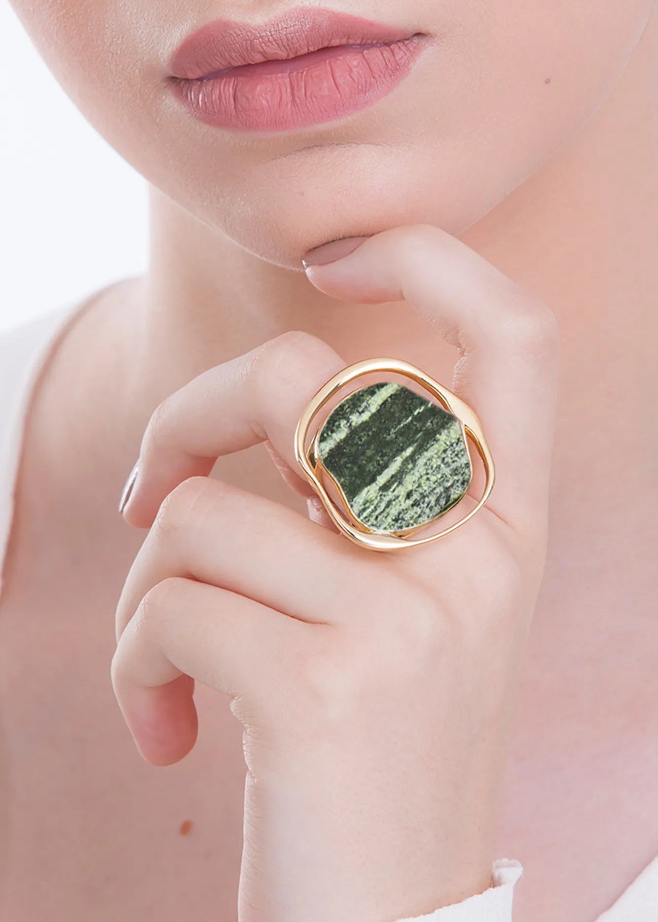 Close-up of a hand wearing a gold ring with a green stone, against a soft pink background.