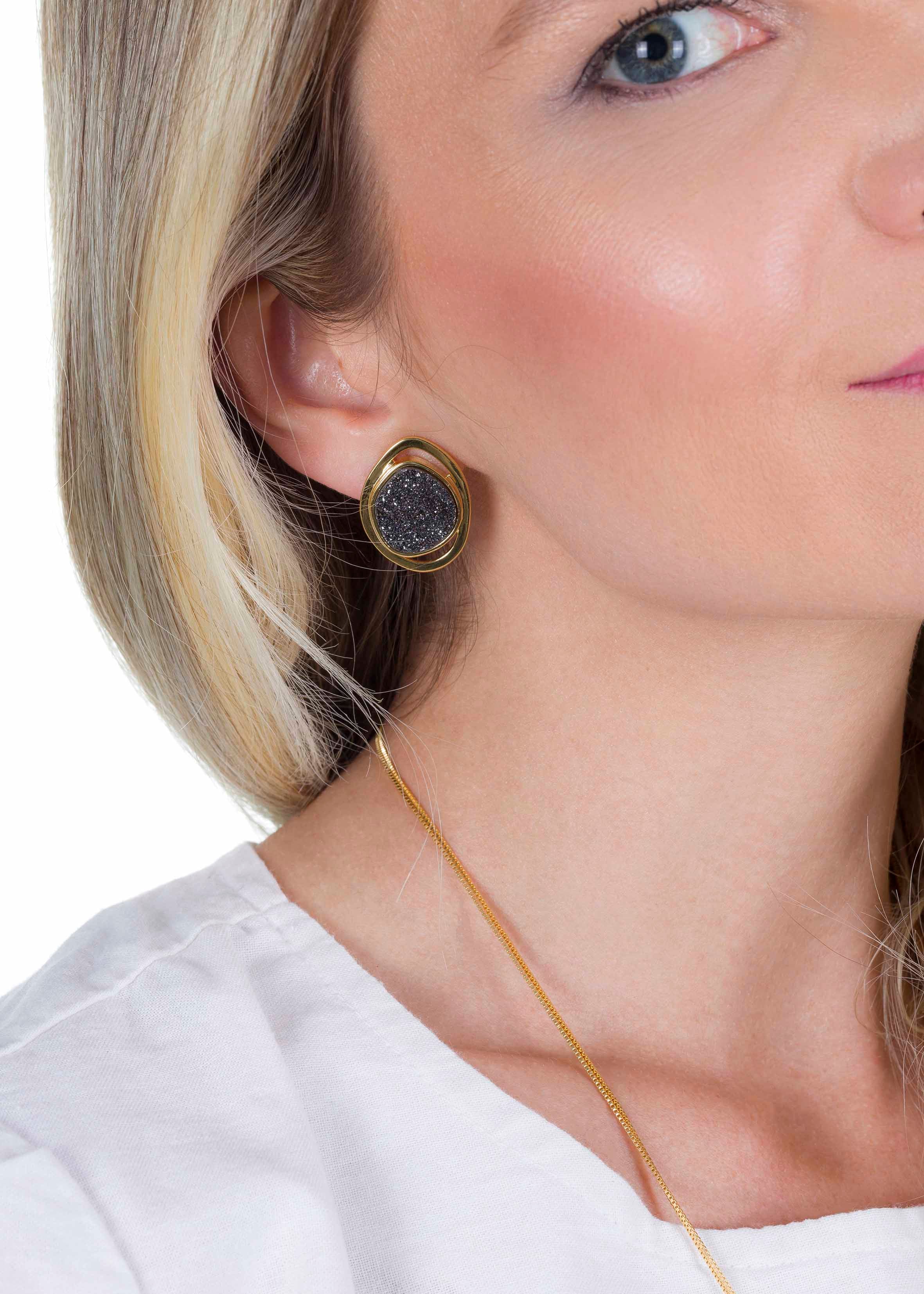 Close-up of a person wearing a gold earring with a dark stone on a plain background