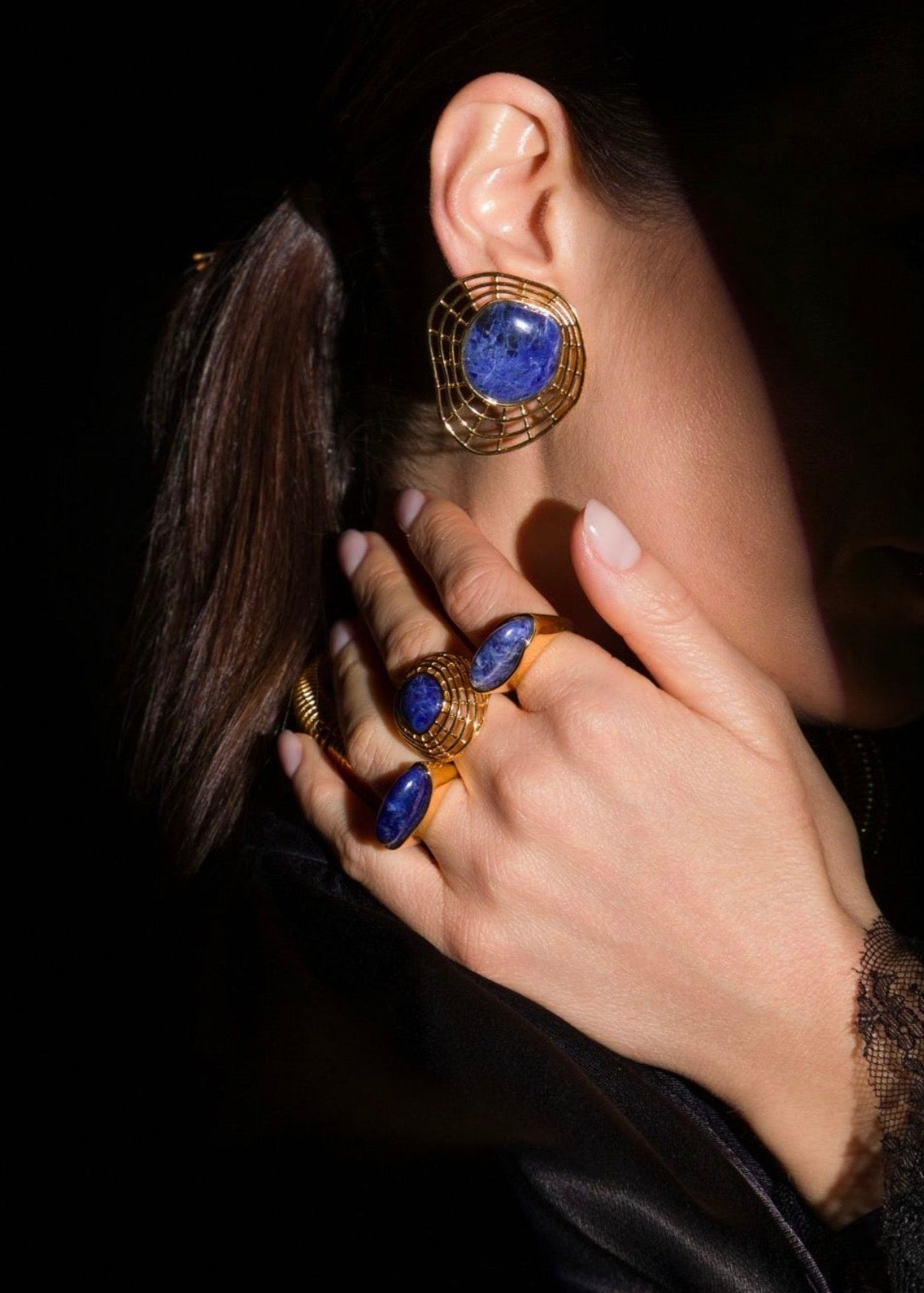 A close-up lifestyle shot of a model wearing gold rings and a matching gold hoop earring with deep blue lapis lazuli stones against a dark background.