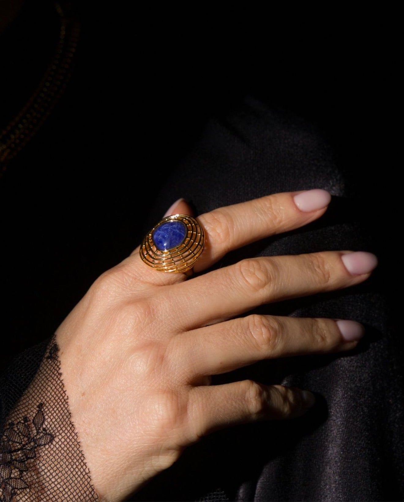 A close-up of a hand wearing a gold ring with a large oval blue stone, set against a dark background.