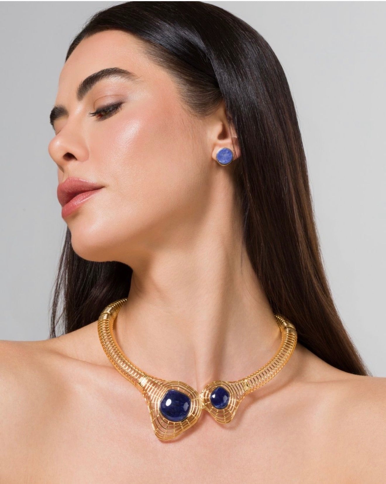 A woman wearing a gold collar necklace with two large oval blue gemstones and a matching blue stud earring, shown against a plain light gray background.