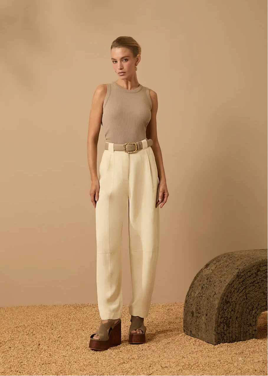 Woman wearing a beige sleeveless top and white pants standing against a beige background