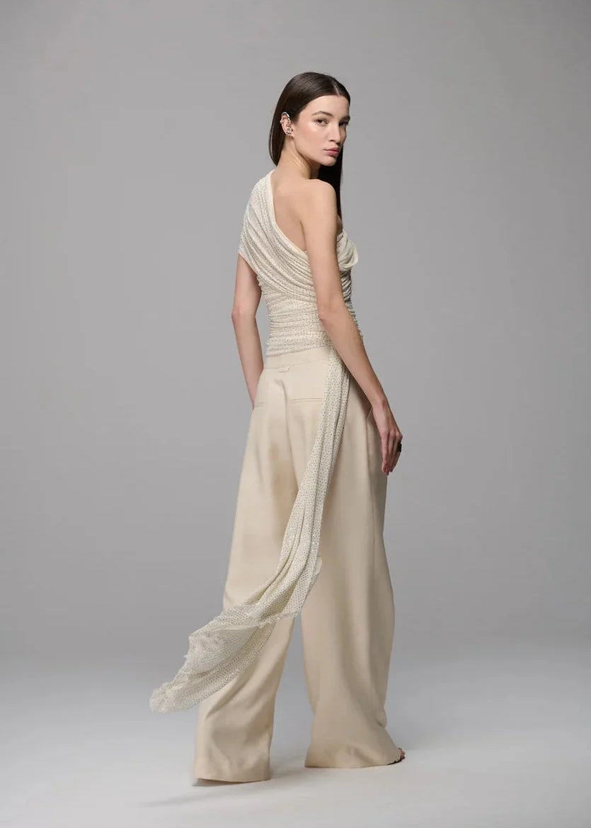 A model wearing a draped, one-shoulder cream-colored gown with wide-leg pants, photographed against a neutral gray studio background.