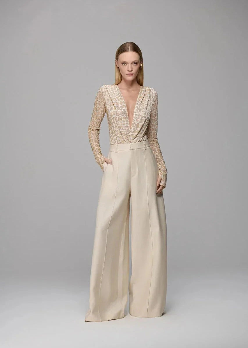 A model stands against a light gray studio background wearing a beige jumpsuit with a heavily textured, patterned deep V-neck bodice and long sleeves, paired with very wide-leg trousers.