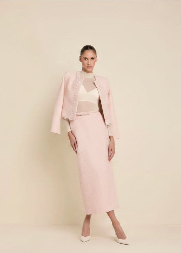 A model stands against a warm beige studio background wearing a coordinated cream outfit consisting of a textured midi skirt, a matching cropped jacket, and a sheer mesh top with white pointed heels.