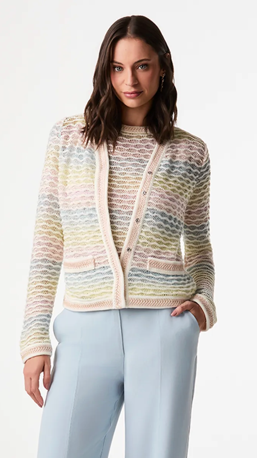 A woman modeling a pastel multicolor knit cardigan with a wavy pattern, paired with light blue trousers, against a plain white studio background.