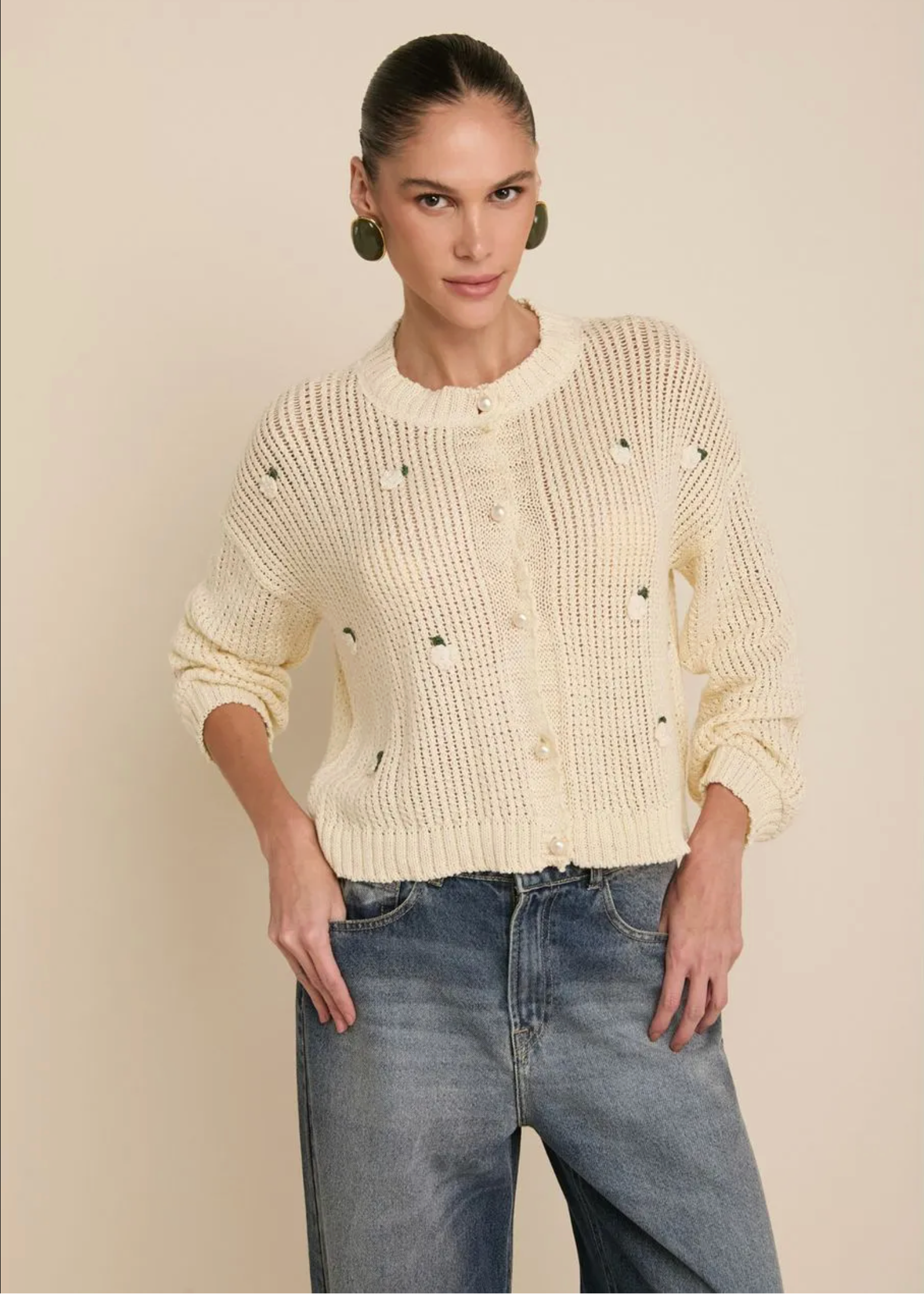 A woman modeling a cream-colored, textured knit cardigan sweater with small embroidered details, paired with blue jeans, against a solid beige background.