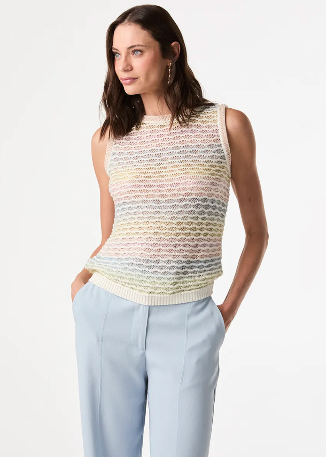 A woman modeling a sleeveless knit top with a multicolor pastel wave pattern, paired with light blue trousers, against a plain white studio background.
