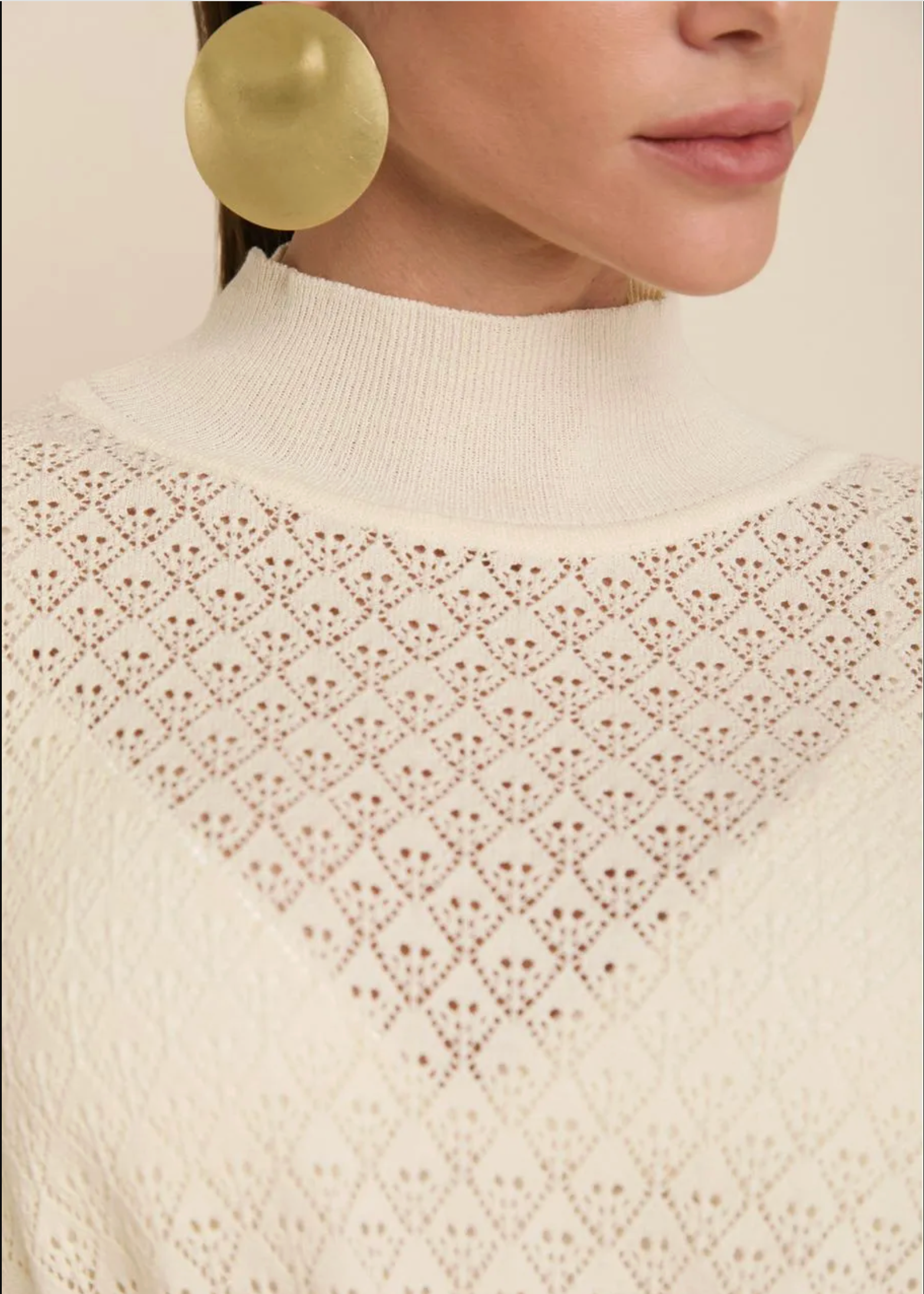 Close-up of a model wearing a cream-colored knit sweater with an intricate openwork diamond pattern and a mock neck, paired with a large gold earring.