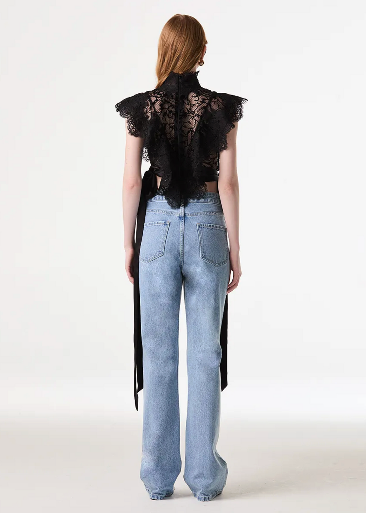 A model viewed from the back wearing a black lace ruffled high-neck top with long tie-back straps and light-wash blue jeans, set against a plain white studio background.