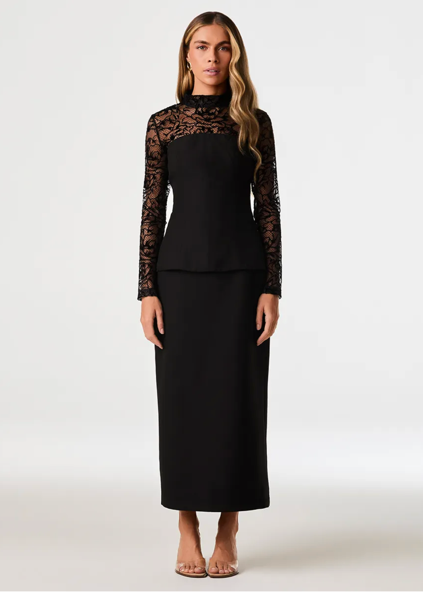 A woman modeling a black two-piece dress with long sheer lace sleeves and a matching midi skirt, photographed against a plain light gray studio background.