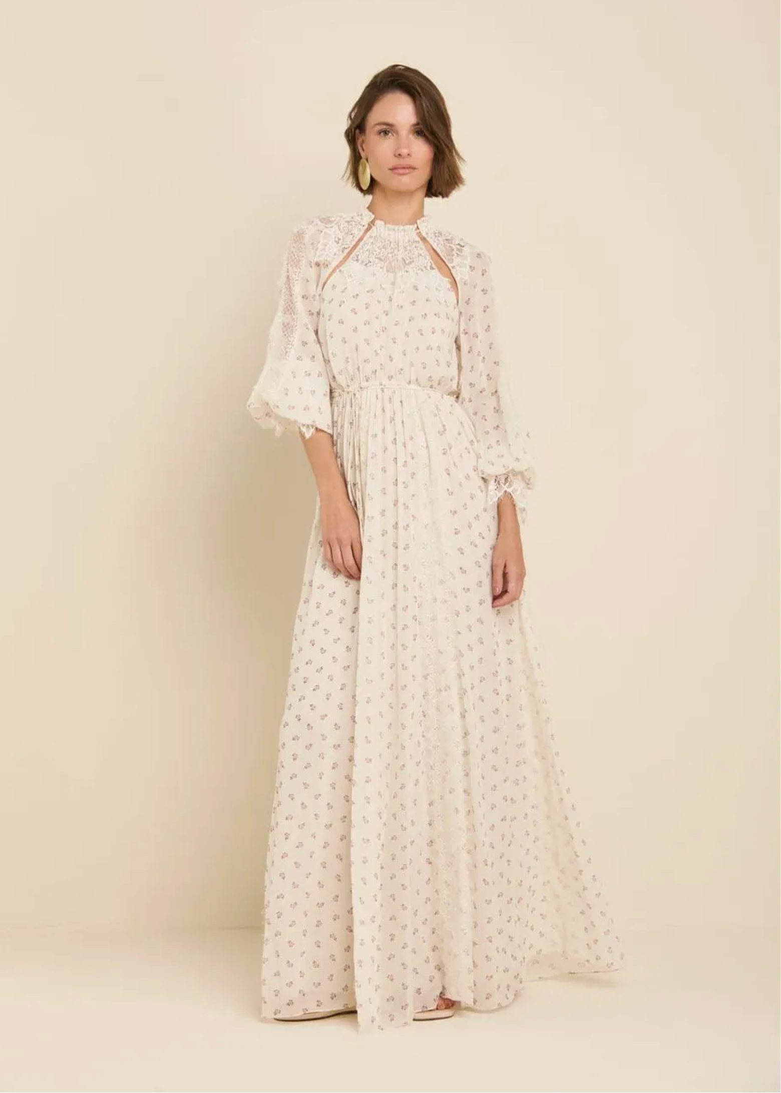 A woman modeling a long cream maxi dress with a small white floral print, featuring a high neckline, sheer elbow-length sleeves, and a gathered waist, set against a solid beige studio background.