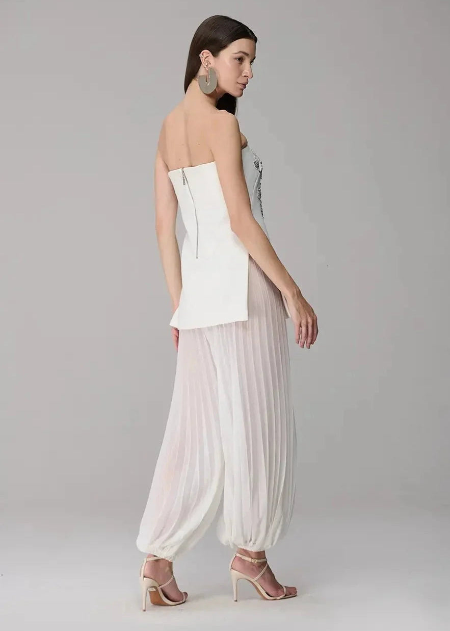 Woman wearing a strapless white top and flowing white pants on a plain background