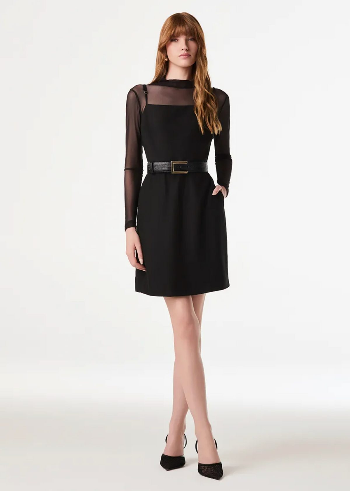 A woman modeling a black long-sleeve shift dress with a sheer mesh yoke and a thin black belt with a gold buckle, paired with black heels, against a plain white studio background.