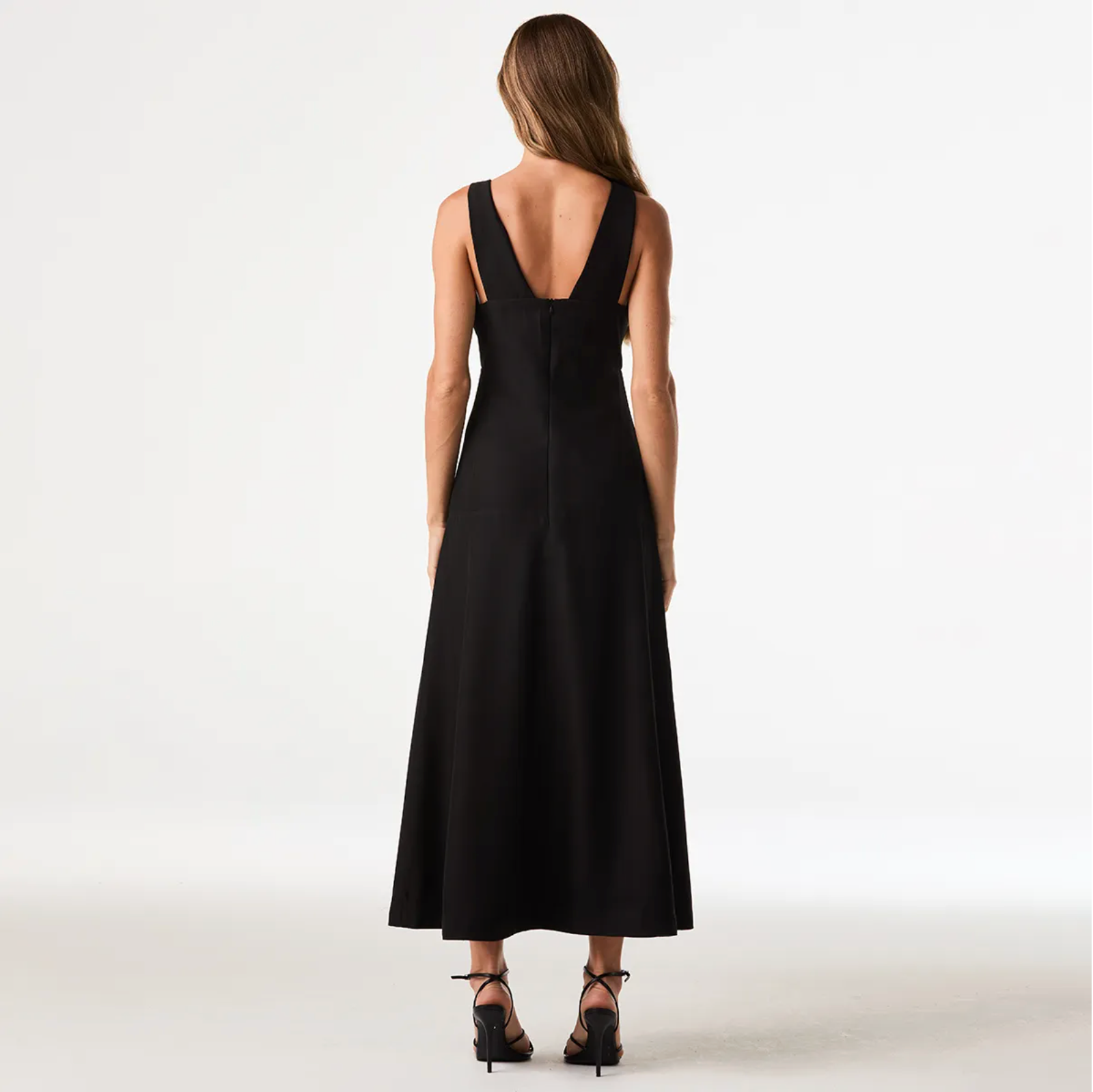 A woman viewed from the back wearing a sleeveless black maxi dress with a deep V-back and black strappy heels, standing against a clean white studio background.