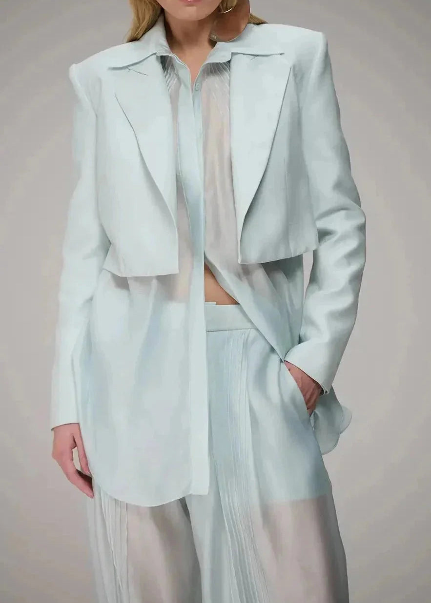 A model wearing a light mint blue tailored jacket with structured shoulders, paired with matching sheer pleated trousers and a sheer inner top, against a neutral studio background.