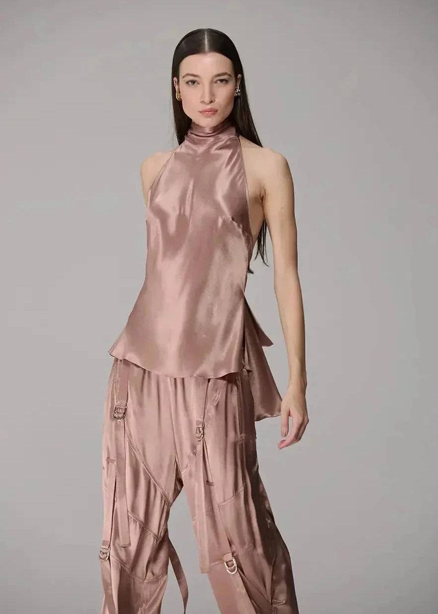 A model wearing a matching two-piece satin outfit in a dusty rose pink color, featuring a sleeveless mock-neck top and wide-leg pants, set against a plain gray studio background.