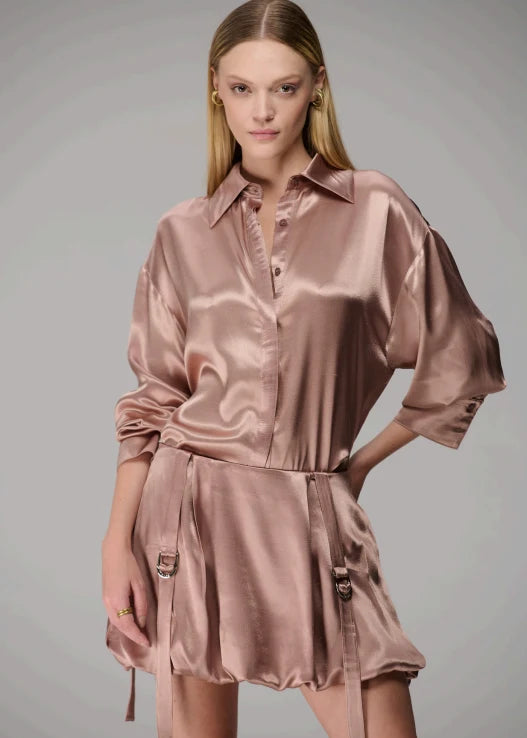 A model wearing a shiny, metallic dusty rose pink satin shirt-jumpsuit with a collar, button front, and a matching mini skirt with side buckle details, set against a plain gray studio background.