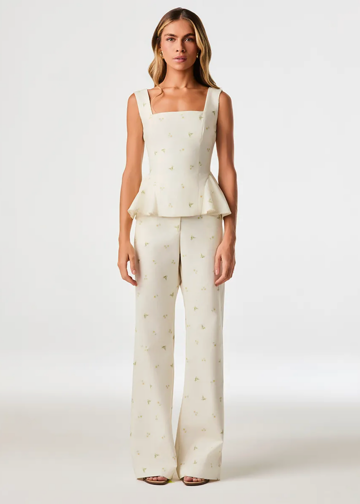 A model stands against a light gray studio background wearing a matching two-piece outfit consisting of a sleeveless square-neck peplum top and wide-leg pants in an off-white fabric with a small gold floral print.