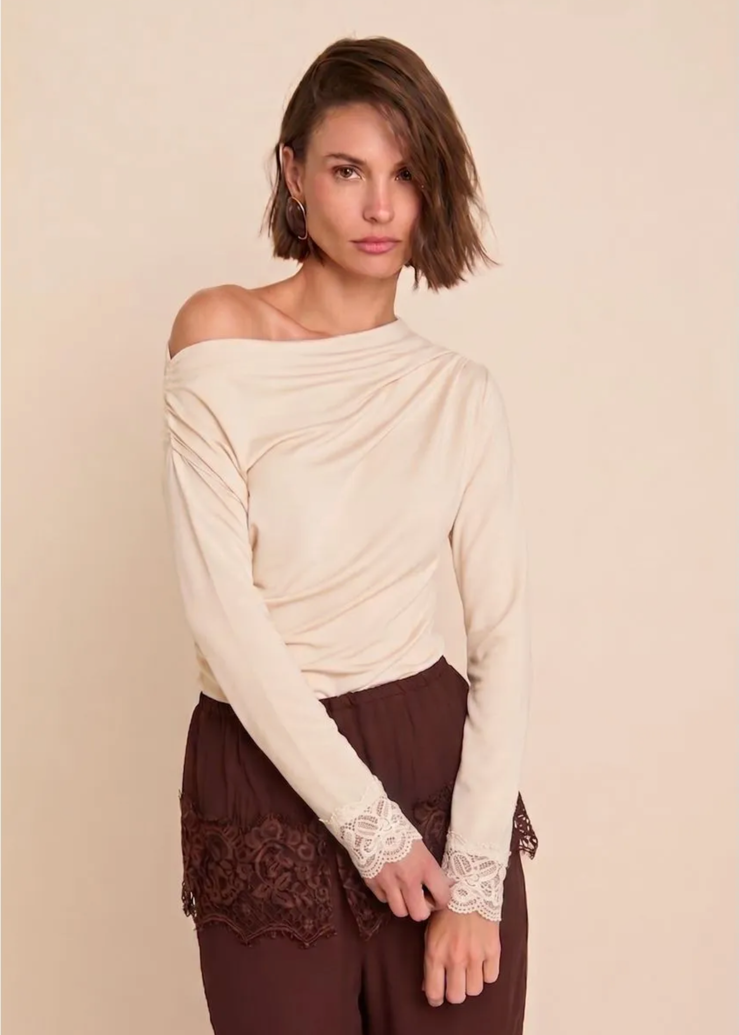 A woman modeling a cream off-the-shoulder long-sleeve top with lace cuffs, paired with dark brown pants featuring lace detailing at the hem, against a solid beige background.