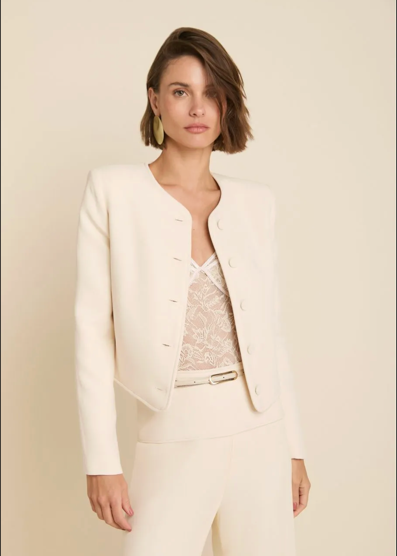 A woman modeling a cream-colored tailored jacket and matching trousers over a lace top, set against a solid light beige background.