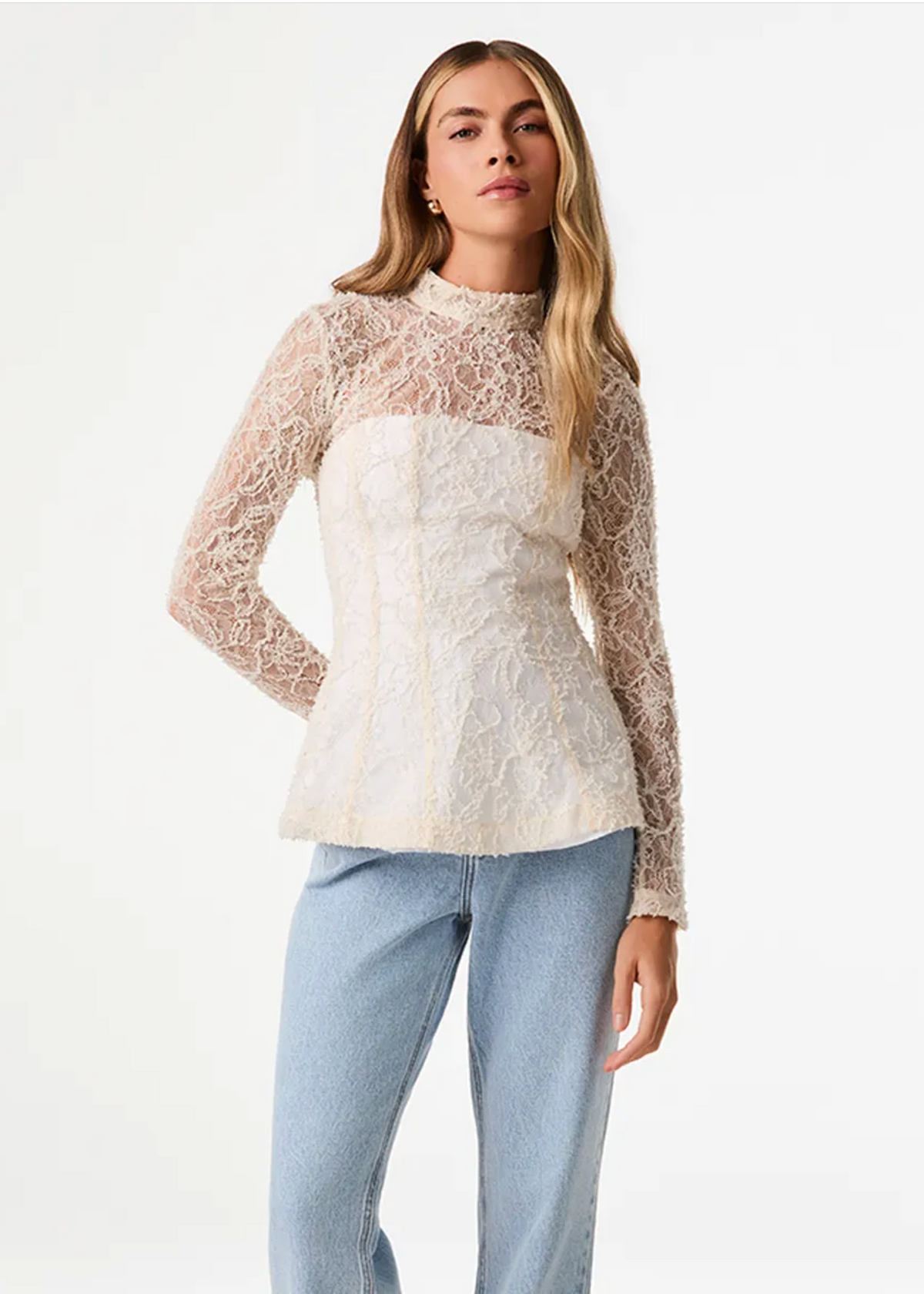 A woman modeling a long-sleeve lace top with a sheer nude lace overlay and a cream lace bust panel, paired with light blue jeans, against a plain white studio background.