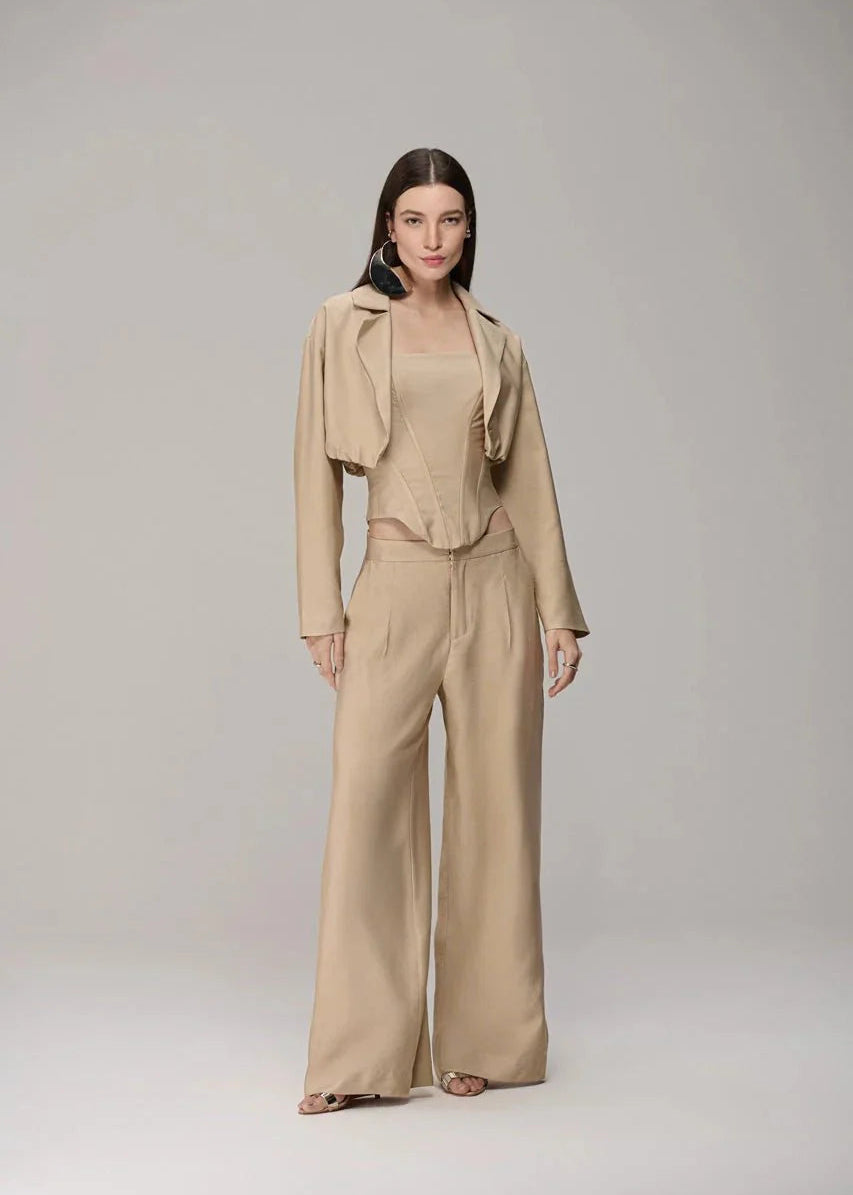 A model stands against a light gray studio background wearing a coordinated beige outfit consisting of a cropped draped jacket, a matching sleeveless top, and wide-leg trousers.