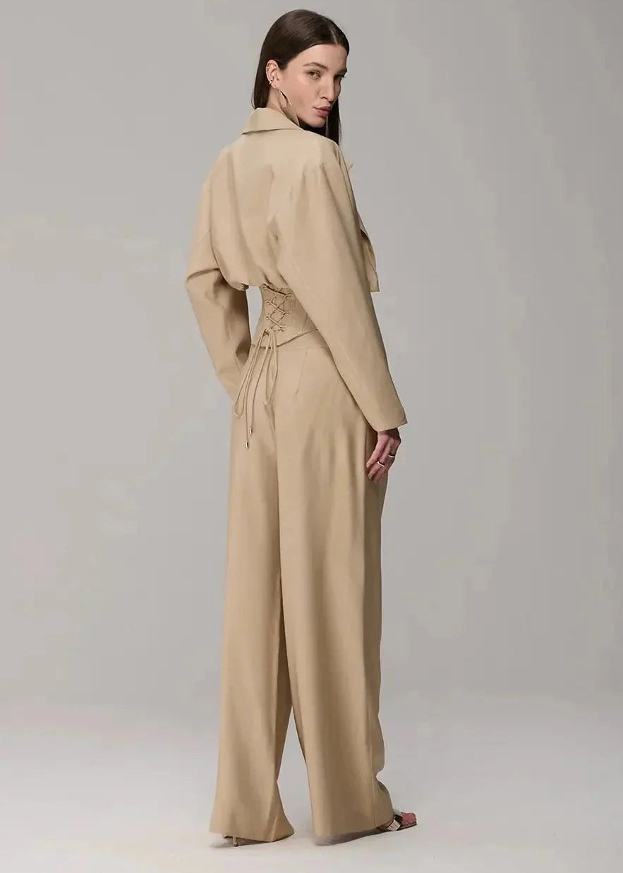 A model is shown from the back wearing a beige wide-leg jumpsuit with a unique draped collar and side tie detail, set against a plain light gray studio background.