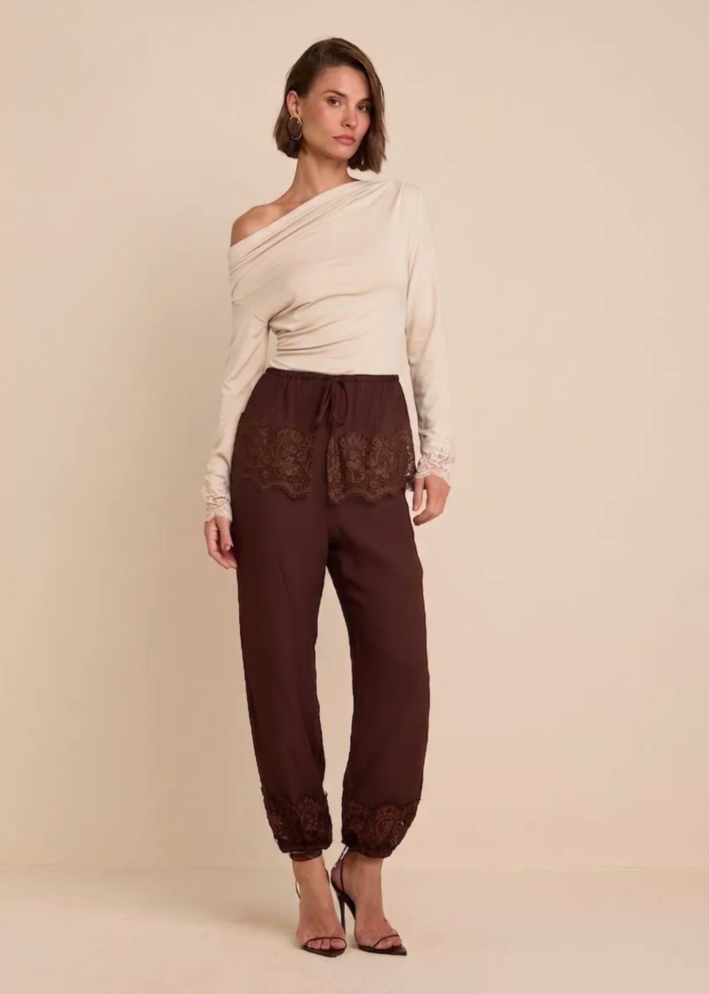 A model stands against a solid beige studio background wearing a cream off-the-shoulder long-sleeve top and dark brown drawstring pants with lace trim at the hips and ankles.