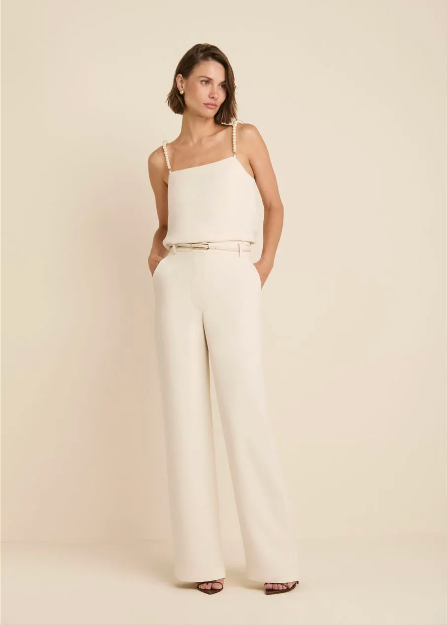 A woman modeling a cream-colored one-shoulder jumpsuit with a thin metallic belt at the waist, photographed against a solid cream studio background.