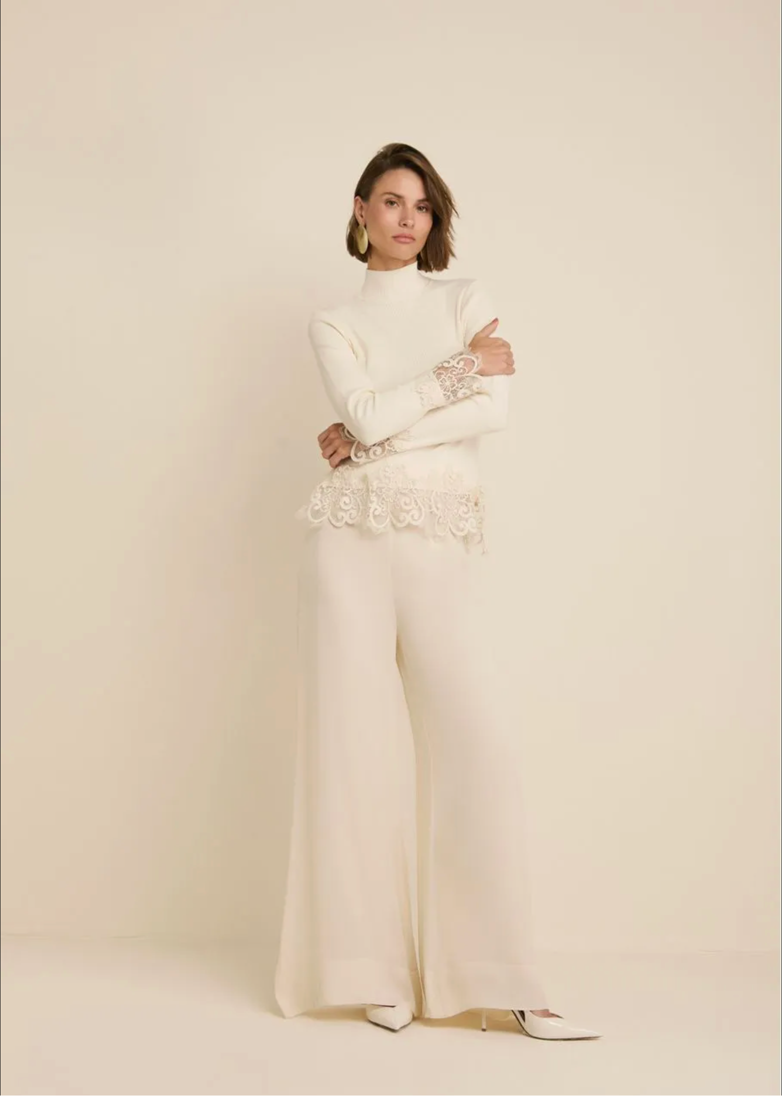 A model stands against a warm cream studio backdrop wearing a matching cream two-piece outfit consisting of a long-sleeve mock-neck top with lace cuffs and a floral lace waistband, paired with wide-leg trousers.
