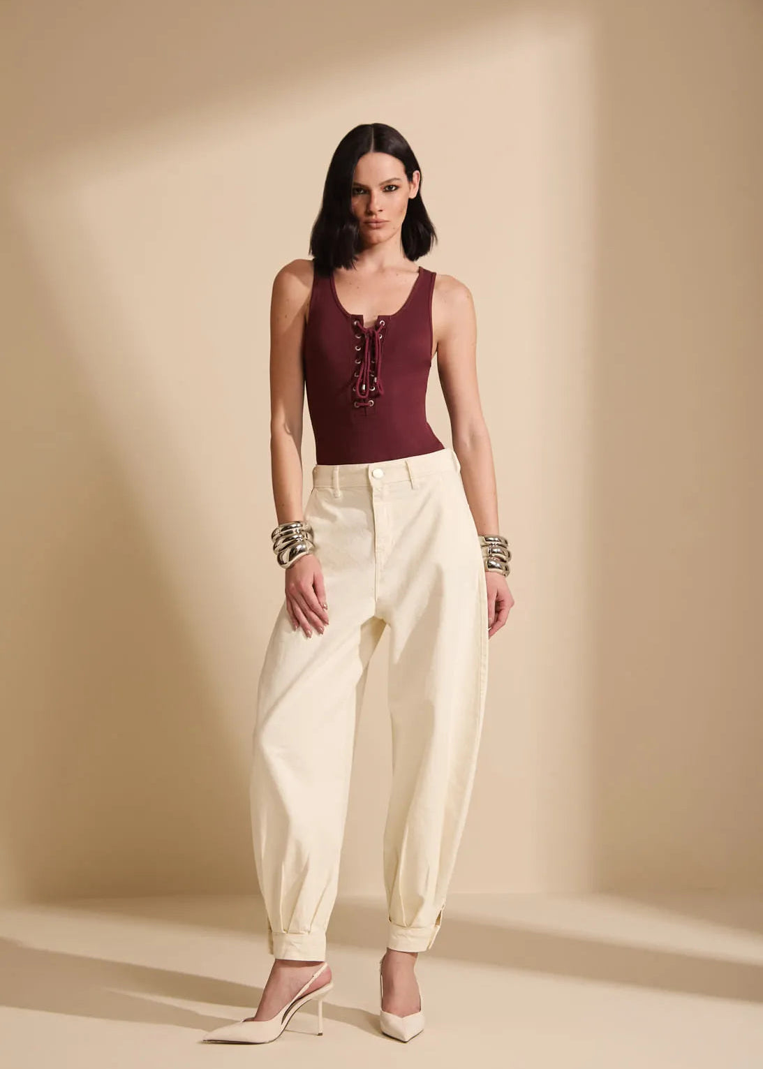 A model stands in a studio wearing a sleeveless burgundy top with lace-up detailing and cream-colored wide-leg cropped pants, paired with white heels and bracelets.