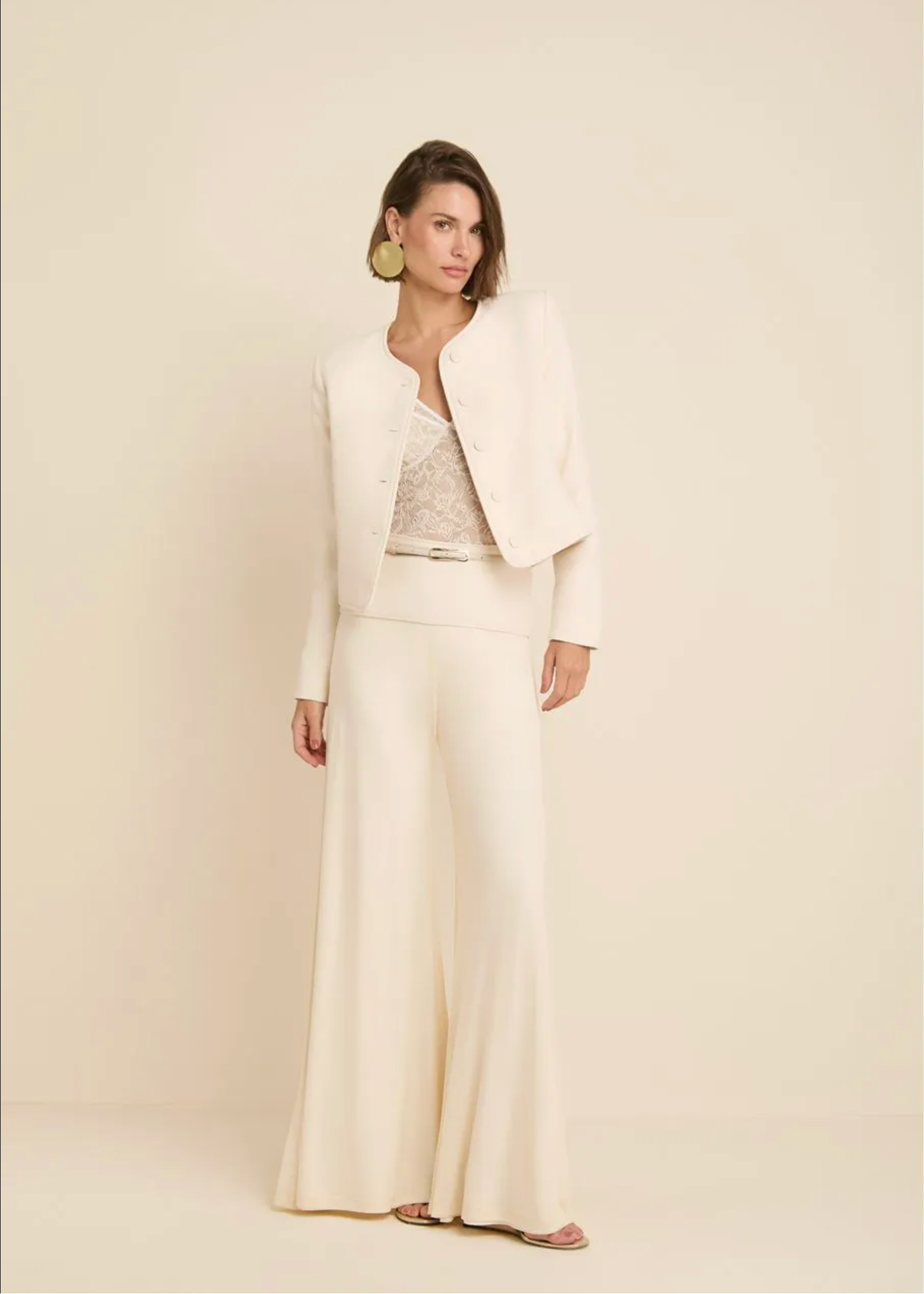 A woman models a cream-colored two-piece suit consisting of a cropped jacket and wide-leg trousers, layered over a lace top, against a solid cream background.