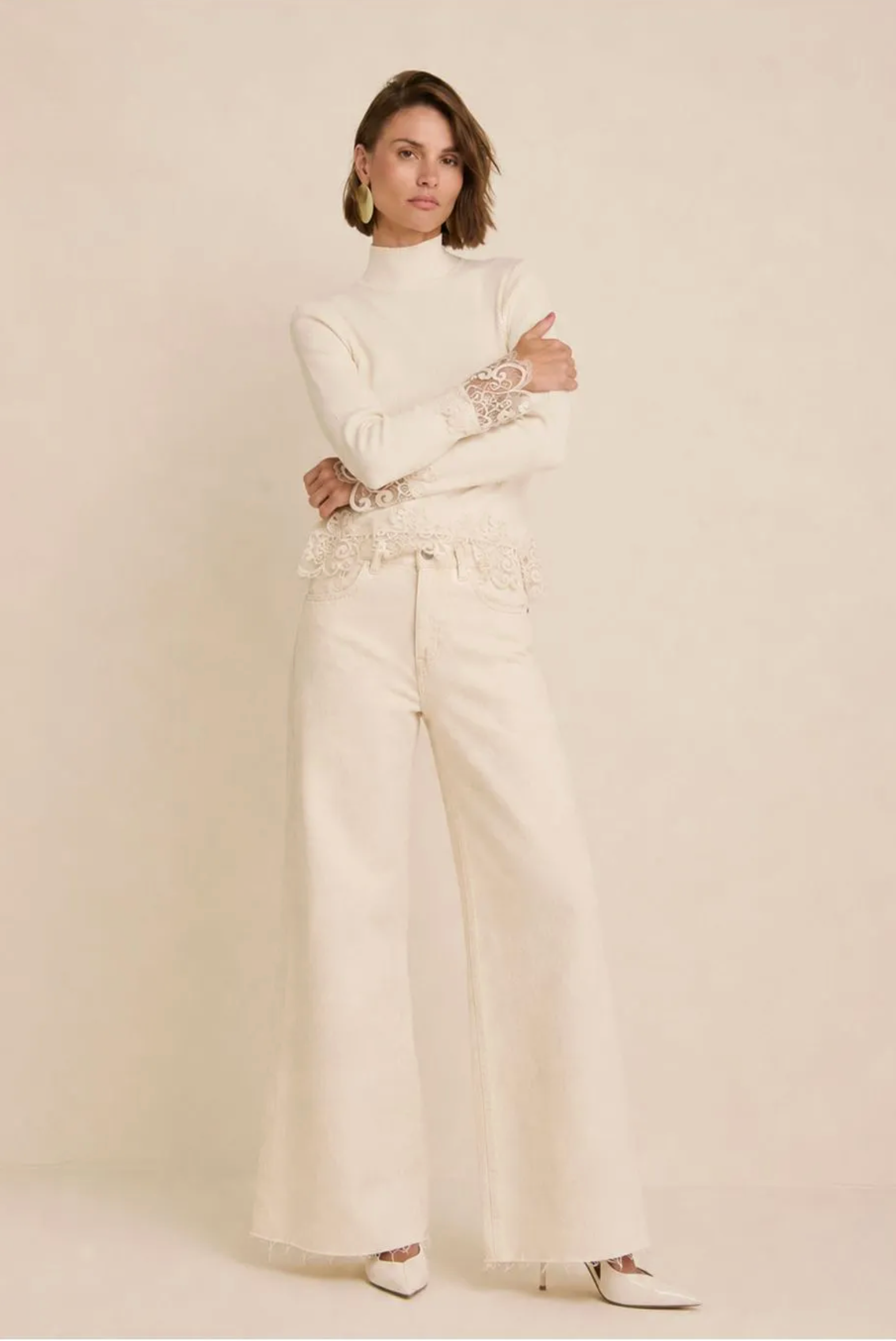 A woman modeling a matching cream-colored two-piece outfit consisting of a long-sleeve mock-neck top with lace detailing on the cuffs and waist, paired with wide-leg trousers, against a solid warm beige studio background.
