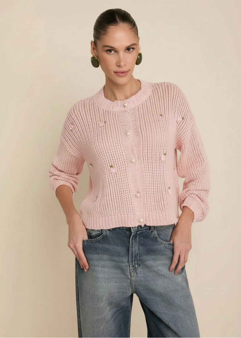 A woman modeling a cream-colored, textured knit cardigan sweater with small embroidered details, paired with blue jeans, against a solid beige background.