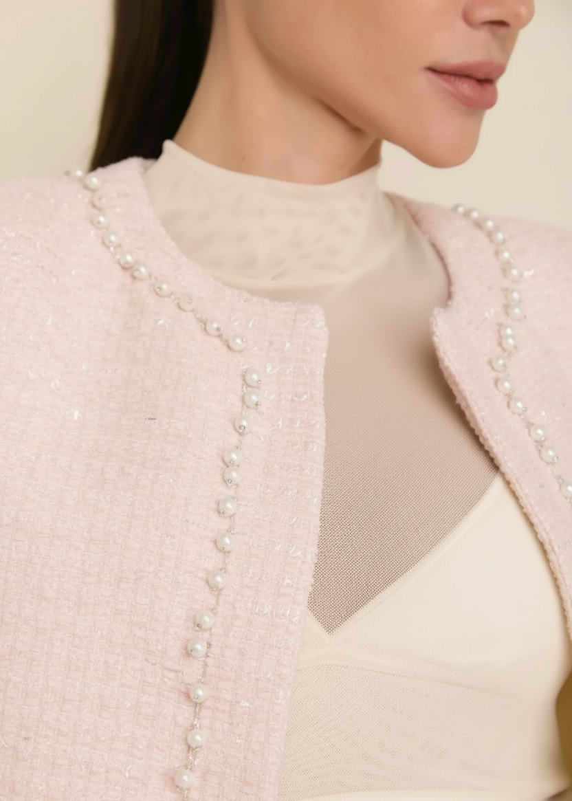 Close-up of a model wearing a cream-colored tweed jacket with pearl embellishments over a sheer cream mock-neck top and bralette.