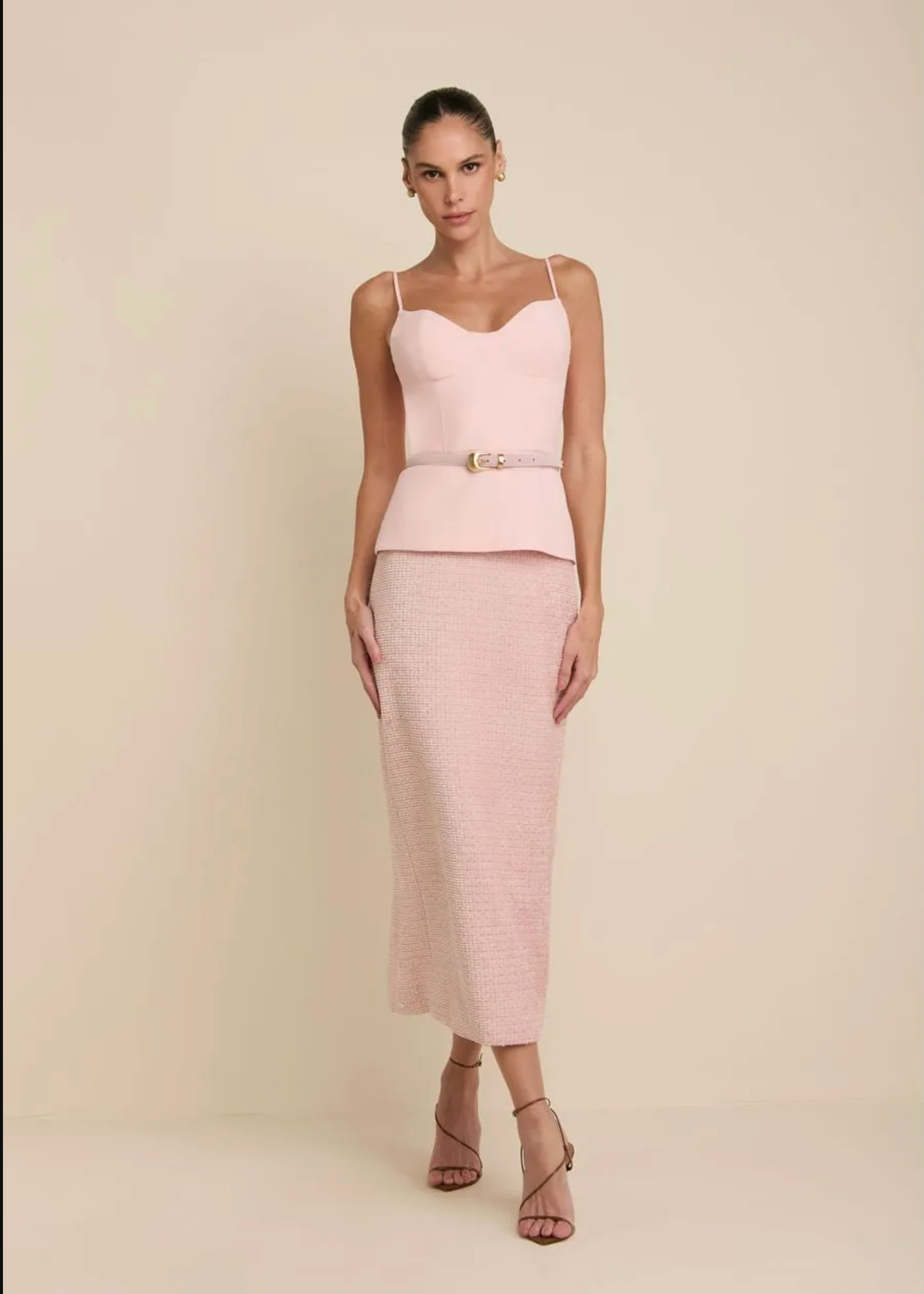 A model wearing a light pink two-piece outfit consisting of a spaghetti-strap peplum top with a matching belt and a textured midi skirt, paired with strappy high-heeled sandals, set against a solid beige studio background.
