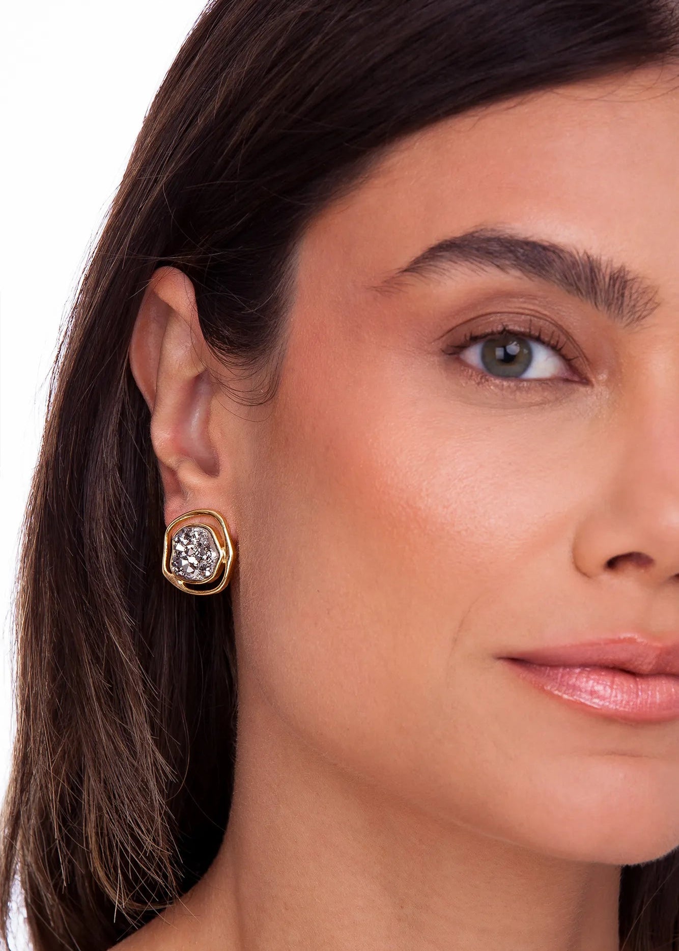 A close-up of a woman's ear wearing a gold-toned geometric stud earring with a clear faceted gemstone, set against a plain white background.