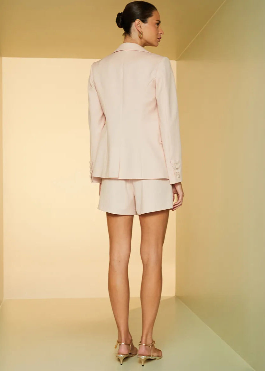 Woman wearing a light pink suit standing in a minimalistic room with soft lighting.