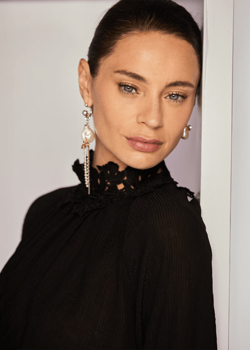 Woman wearing a black top with floral details and pearl earrings against a light background
