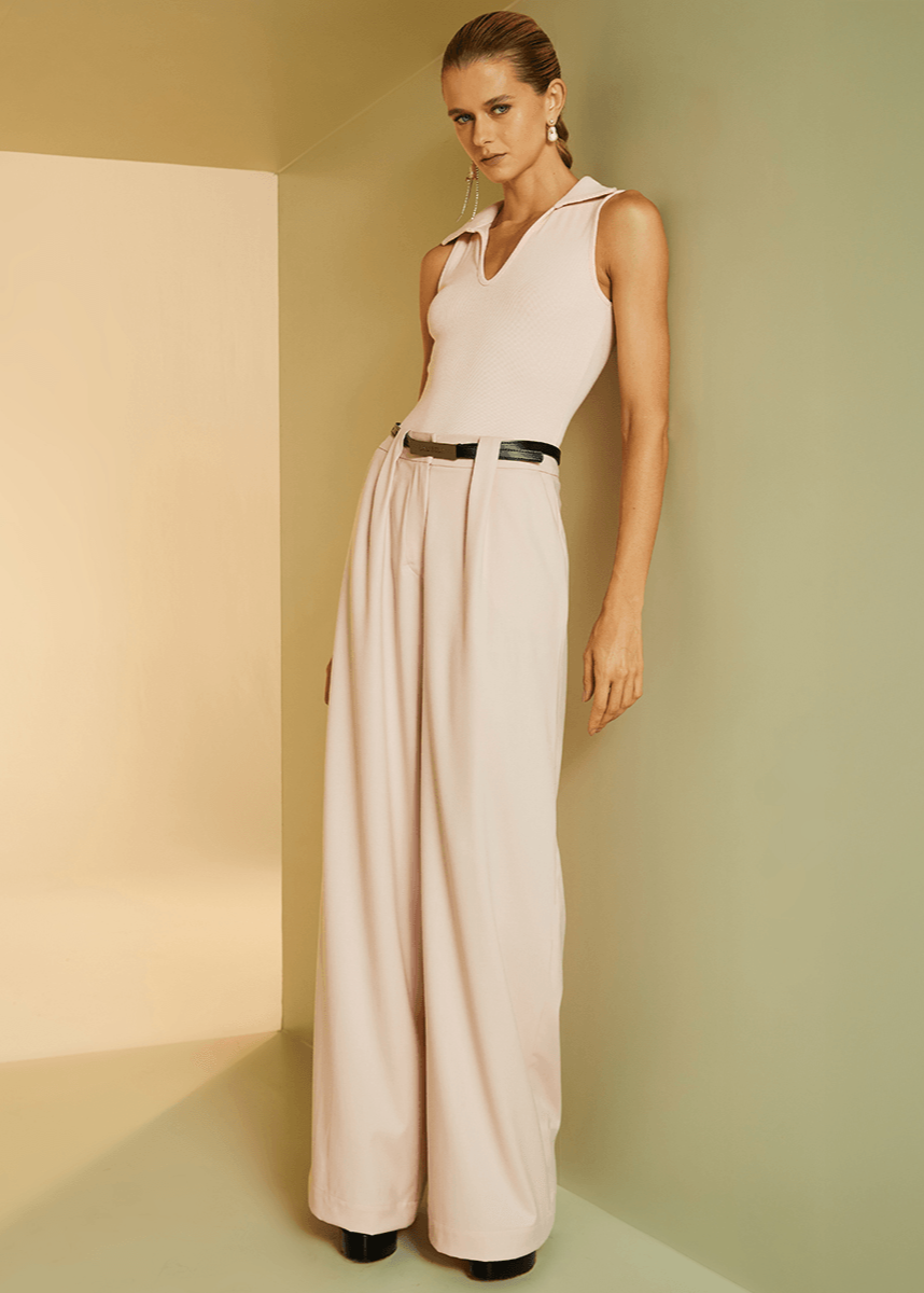 Woman wearing a light pink sleeveless jumpsuit with a belt against a beige wall.