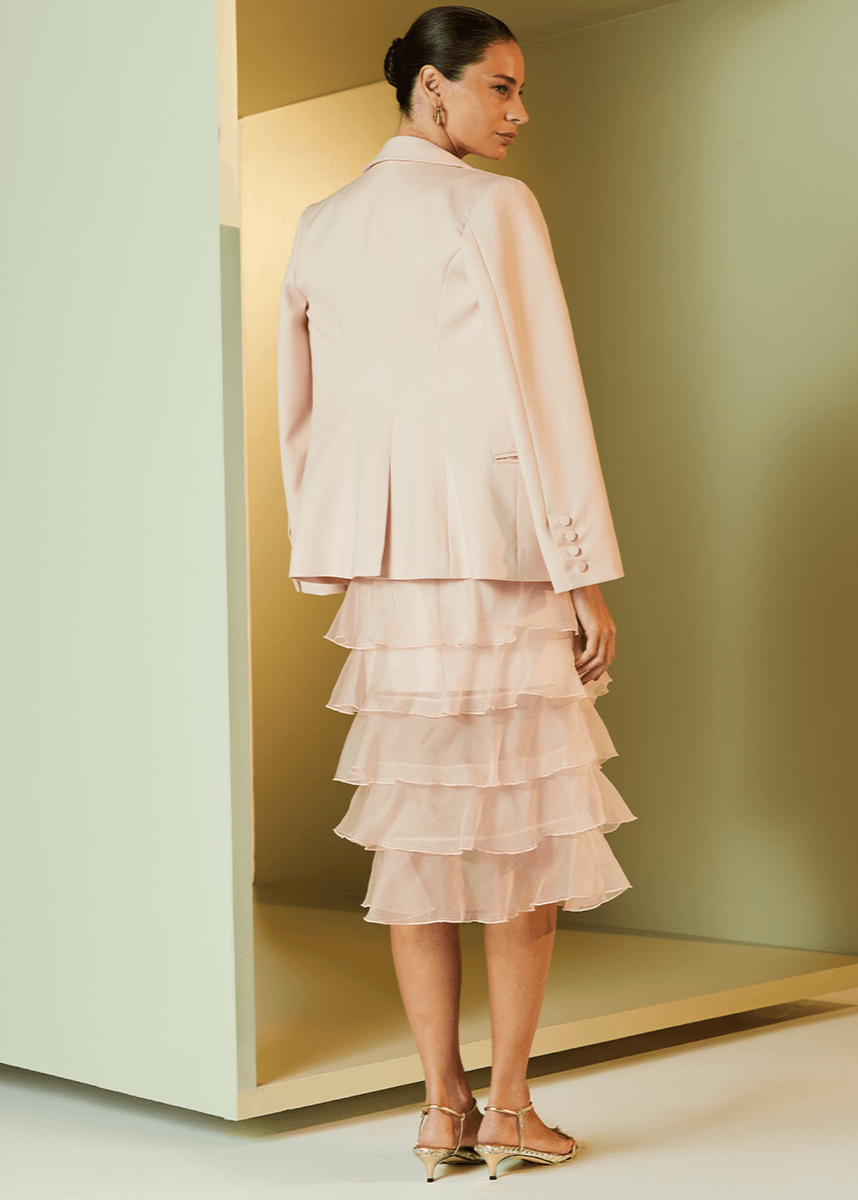 Woman wearing a light pink ruffled dress and jacket in a minimalistic setting.