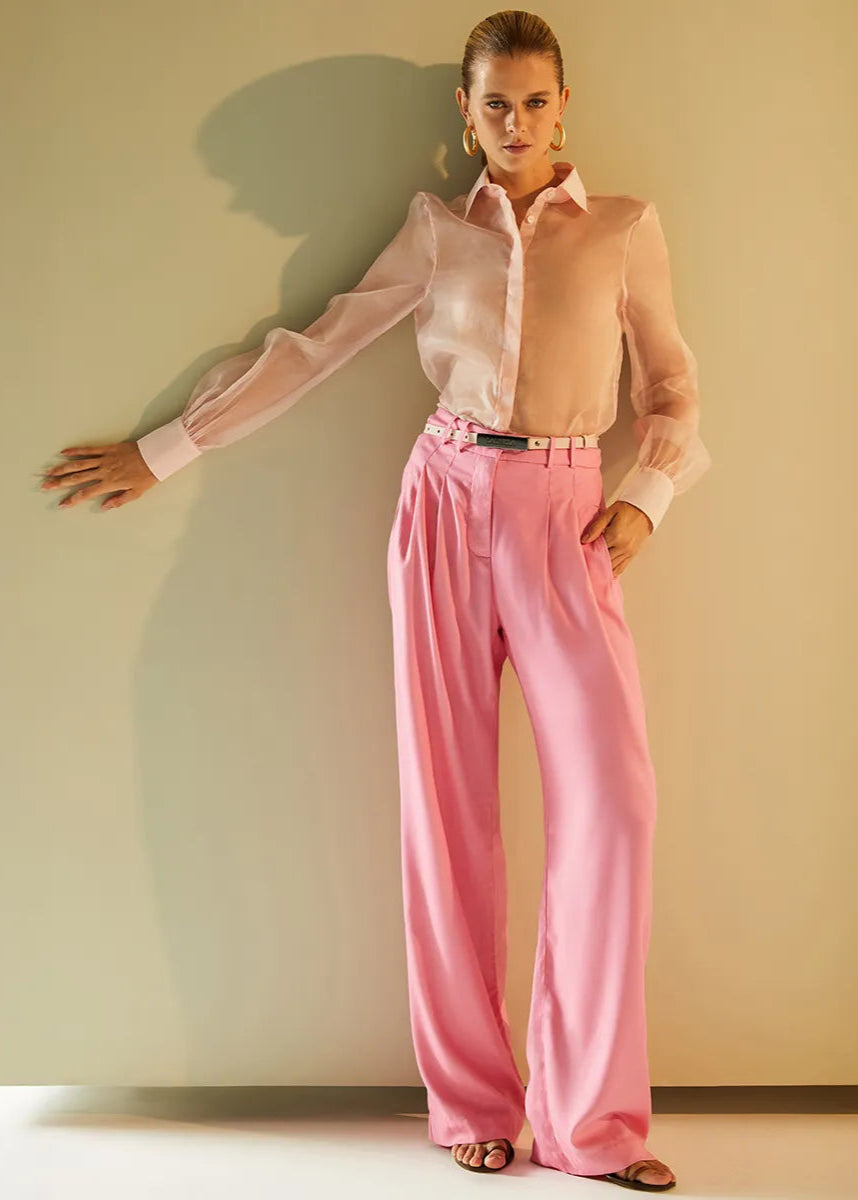 Woman wearing a light pink shirt and pink pants against a plain wall.
