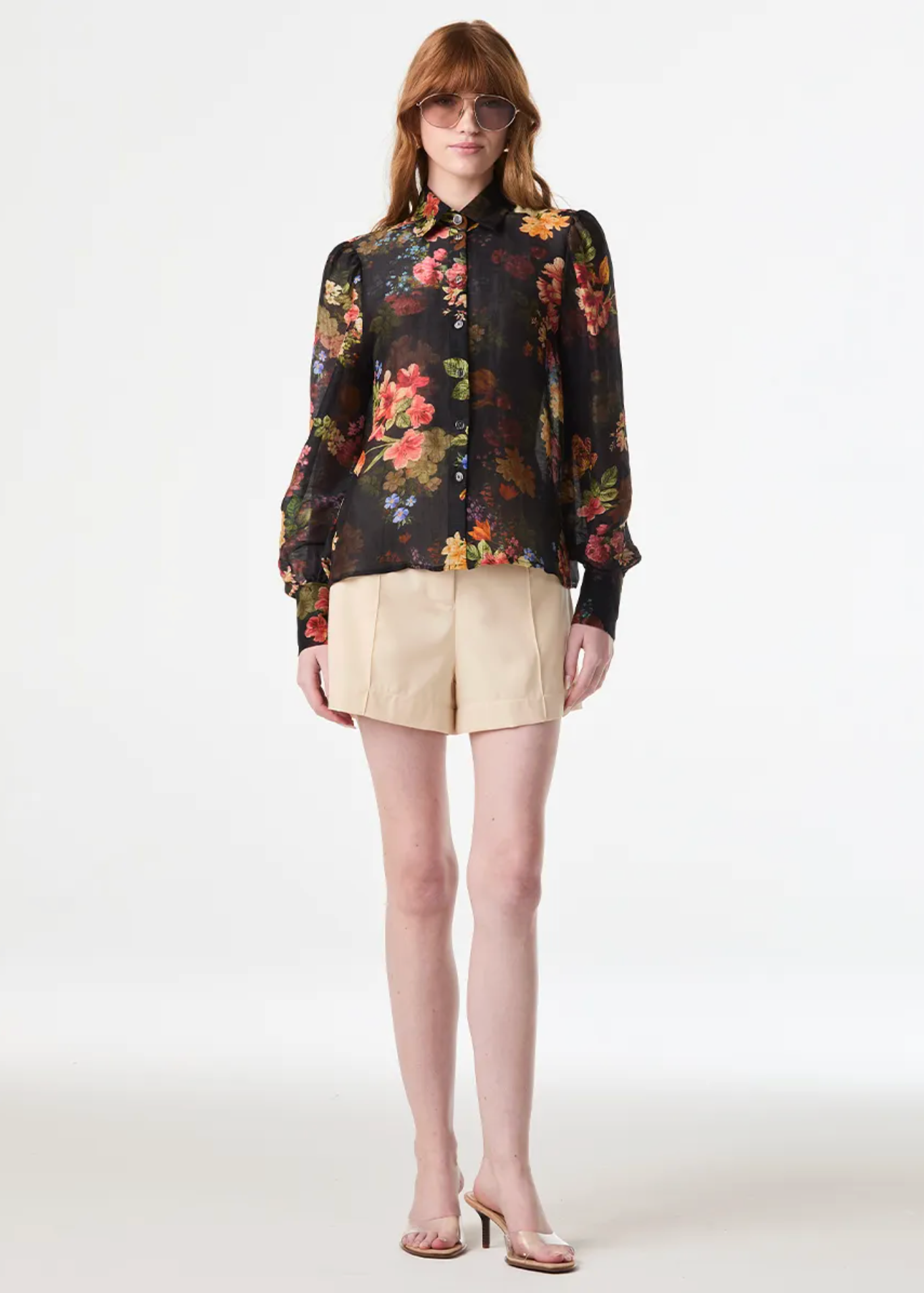 A model stands against a plain light gray studio background wearing a black long-sleeve button-up blouse with a dense multicolor floral print, paired with beige shorts and clear high-heel sandals.
