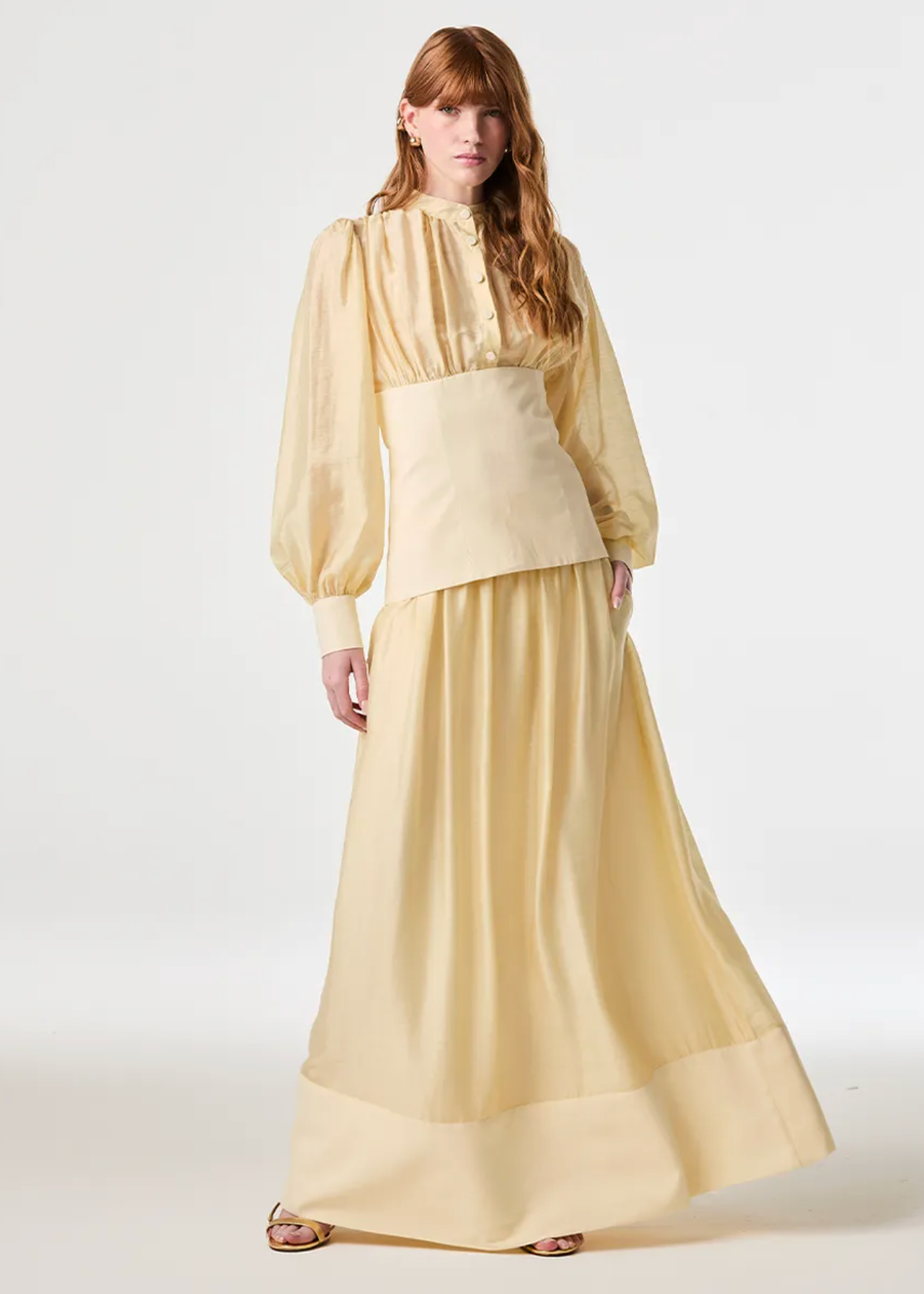 A woman modeling a pale yellow, long-sleeved maxi dress with a structured waist and a wide hem, photographed against a seamless white studio background.