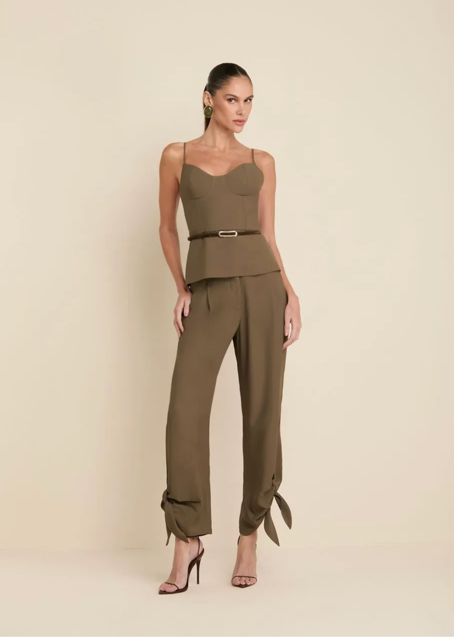 A model wears an olive green sleeveless jumpsuit with a structured sweetheart bodice, a matching belt with a silver buckle, and wide-leg cropped pants with tie details at the ankles, set against a solid beige studio background.